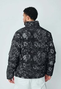 Veste matelassée noire avec un motif floral dans des nuances de gris. Elle possède un col montant et une texture matelassée.