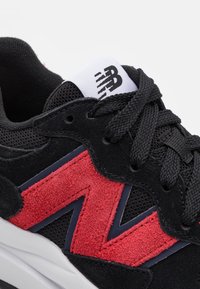 Musta urheilukenkä, jossa on punaisia mokka-yksityiskohtia ja verkko-osia. Valkoinen brändäys kengän kielessä, litteät mustat nauhat ja pehmustettu pohja.