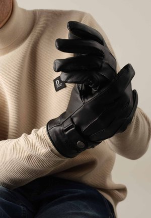 Gants en cuir noir avec une texture lisse, dotés de doigts articulés et d'une sangle sécurisée avec quincaillerie en métal, portés par une personne en pull beige.
