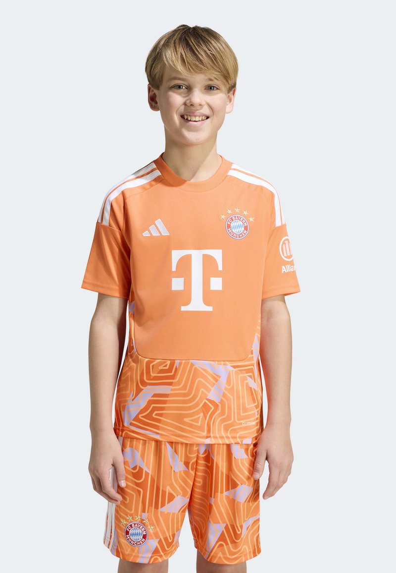 Lächelnder Junge, der ein orangefarbenes FC Bayern München Fußballtrikot und Shorts mit weißen und violetten abstrakten Mustern trägt, steht vor einem schlichten Hintergrund.