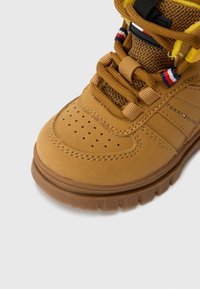 Sneaker en suede marron avec des détails perforés, des accents en maille et des lacets jaunes. Une marque notable est présente sur le côté. Semelle en caoutchouc épaisse.