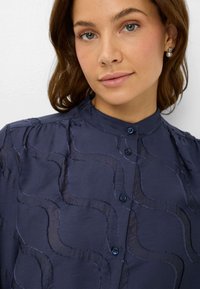 Blusa blu navy con bottoni, colletto alto, caratterizzata da un motivo ondulato testurizzato, tessuto delicato e bottoni blu, fotografata dal torace in su.