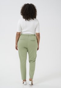 Pantalons verts à coupe décontractée avec une taille élastique, deux poches arrière et une texture lisse, associés à un haut blanc et des chaussures légères.