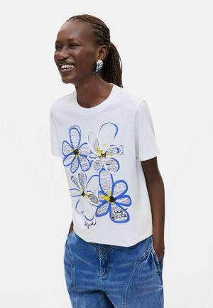 Frau mit geflochtenem Haar lacht, trägt ein weißes T-Shirt mit großen blauen und weißen Blumen und blaue Jeans, die Hände in den Taschen.