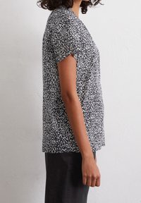 Femme portant un haut à manches courtes noir et blanc à motif, ainsi qu'une jupe noire, debout de profil contre un mur blanc uni.