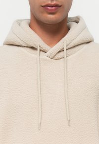 Beige Fleece-Hoodie mit einer weichen Textur, mit Kordelzug und einer Kängurutasche. Einfaches Design, keine sichtbaren Muster oder Hardwaredetails.