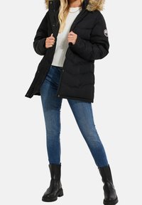 Kvinde iført en sort pufferjakke med pelselv, hvid ribbet sweater, blå skinny jeans og sorte ankelstøvler, der står foran en hvid baggrund.