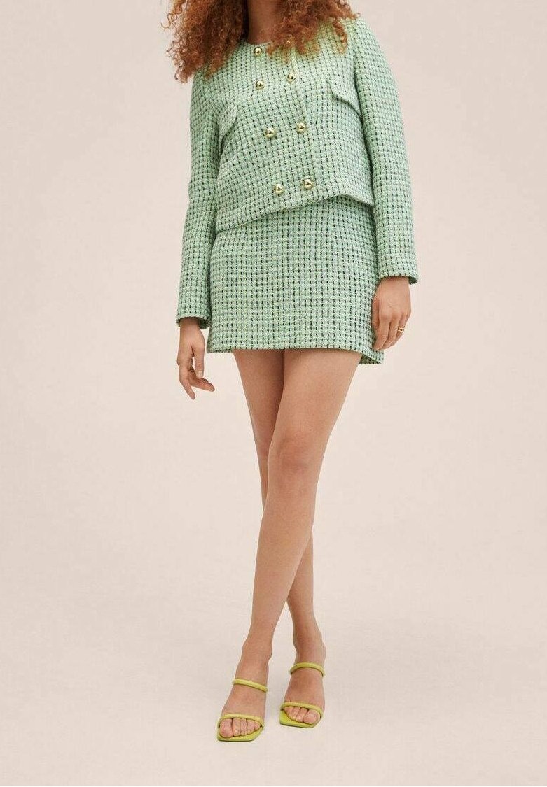 Femme portant une veste en tweed vert clair avec des boutons dorés et une mini-jupe assortie, accompagnée de talons à lanières vert citron.