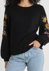 Black long-sleeve sweatshirt with embroidered yellow and red flowers on sleeves, worn with light blue jeans.