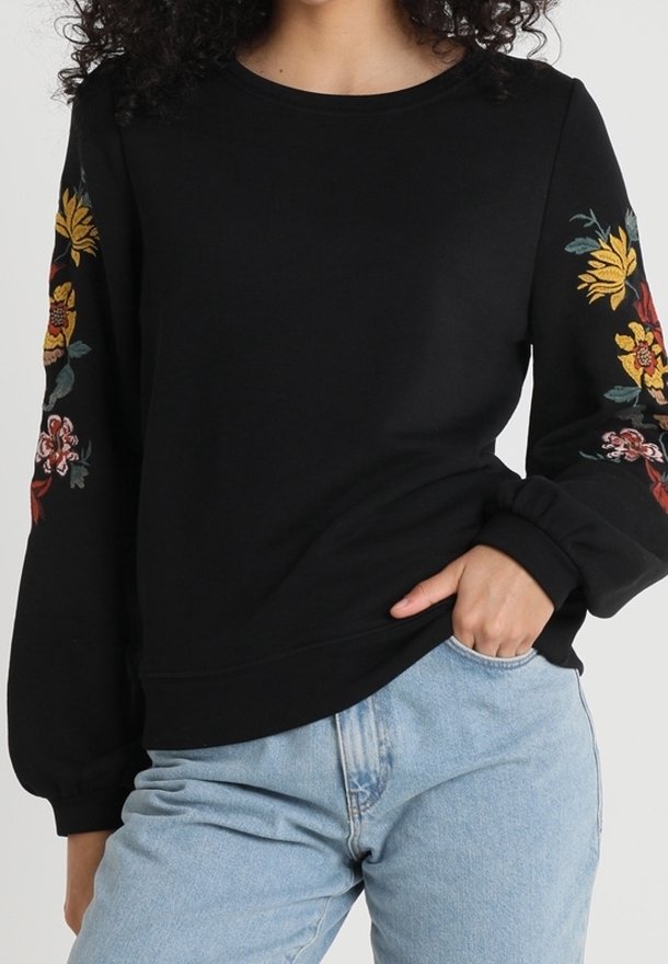Black long-sleeve sweatshirt with embroidered yellow and red flowers on sleeves, worn with light blue jeans.