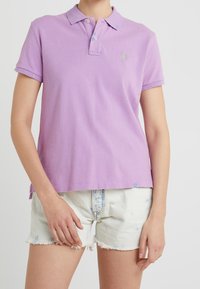 Lilacová polo tričko s límečkem, krátkými rukávy a malým zeleným logem. Nosené s bílými, opotřebovanými denimovými šortkami.