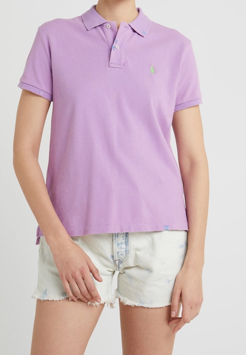 Lilacová polo tričko s límečkem, krátkými rukávy a malým zeleným logem. Nosené s bílými, opotřebovanými denimovými šortkami.