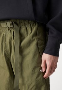 Handen vilar bredvid olivgrön byxor med elastisk midja och bälte, tillsammans med en svart långärmad tröja.