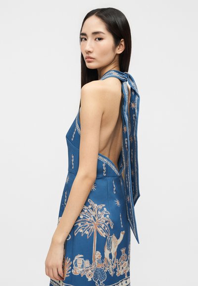 Νεαρή γυναίκα με μακριά σκούρα μαλλιά, φορώντας μπλε φόρεμα με λαιμόκοψη halter, χωρίς πλάτη και διακοσμητικό σχέδιο με φοινικόδεντρα και καμήλες.