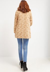 Tanfärgad faux fur-kappa med ett strukturerat, cirkulärt mönster, långa ärmar och en knälång design, kombinerad med blåa skinny jeans och svarta ankelstövlar.