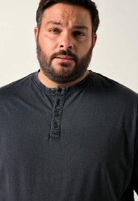 Vousatý muž s krátkými tmavými vlasy, oblečený v černém tričku henley s čtyřmi knoflíky, na jednoduchém světlém pozadí.