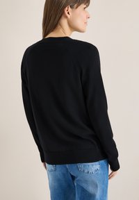 Femme aux cheveux bruns portant un pull noir à manches longues et un jean bleu, vue de dos, debout devant un fond uni.