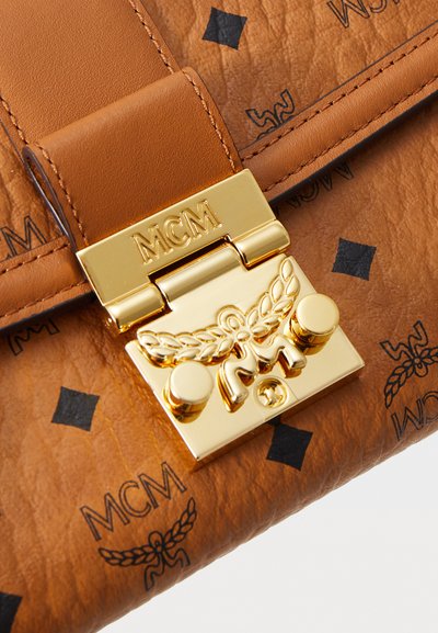 Karamelist nahkakott, millel on kuldne kinniti, millel on graveeritud "MCM" ja mis on kaunistatud tiibade embleemiga, sisaldades mustast teemantist ja logomustriga nahka.