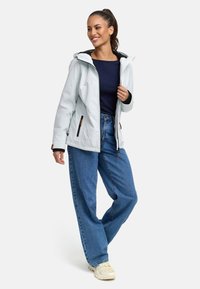Veste imperméable bleu clair avec capuche, doublure noire, poches zippées, associée à un jean bleu ample et des baskets beiges.