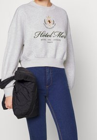 Sweatshirt gris avec texte brodé, poignets et ourlet côtelés, associé à un jean bleu taille haute ; un sac noir rembourré est porté sur le bras.