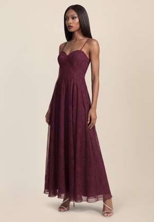 Vrouw die een vloerlang, mouwloos bordeaux kanten jurk met dunne bandjes en zilveren hoge hak sandalen draagt, staand tegen een effen achtergrond.