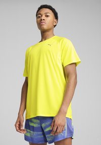 Gul sports t-shirt med korte ærmer, lavet af letvægtsstof. Har et Puma-logo på brystet. Kombineret med blå shorts med et struktureret mønster.