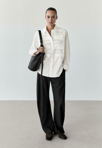 Bílá košile s knoflíky s texturovanými sklady na hrudi, doplněná černými kalhotami s širokými nohavicemi a černou texturovanou kabelkou přes rameno.