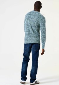 Man die met zijn rug naar de kijker staat, gekleed in een blauwgroene gebreide trui, donkerblauwe jeans en witte sneakers met zwarte strepen op een effen witte achtergrond.