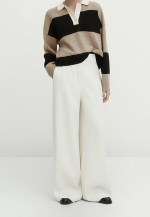Persoon die een beige en zwart gestreepte polo trui draagt met wijde witte broekspijpen en zwarte schoenen, staand tegen een effen achtergrond.