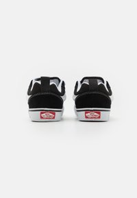 Sneakers suede negre și albe cu talpă joasă, călcâi întărit, talpă intermediară albă și branding roșu pe spate. Suprafață texturată, design minimalist.