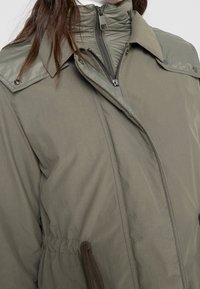 Olivgrön vadderad jacka med hög krage, dragkedja, tryckknappar och justerbar dragsko i midjan. Slätt, vattenavvisande tyg.