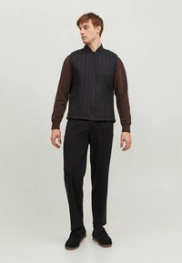 Schwarze Steppweste mit vertikaler Steppung, Reißverschluss und Stehkragen; kombiniert mit braunem Langarmshirt und schwarzen Hosen.