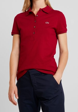 Kobieta ubrana w czerwoną koszulkę polo z krótkim rękawem i guzikami oraz granatowe spodnie, stoi z jedną ręką w kieszeni na szarym tle.