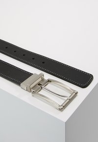 Cintura in pelle nera con cuciture bianche, dotata di una fibbia in metallo argento e fori regolabili per la chiusura. Cintura posizionata su una superficie bianca.