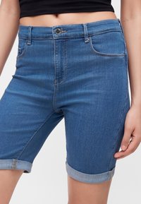 Primer plano de una persona con pantalones cortos vaqueros azules de media pierna con bajos vueltos y un top corto negro, con la mano apoyada al lado del cuerpo.