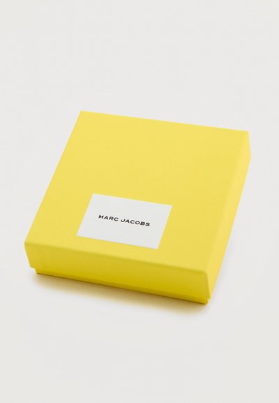 Φωτεινό κίτρινο τετράγωνο κουτί με λευκή ετικέτα που εμφανίζει το "MARC JACOBS" με μαύρο κείμενο σε ανοιχτό φόντο.