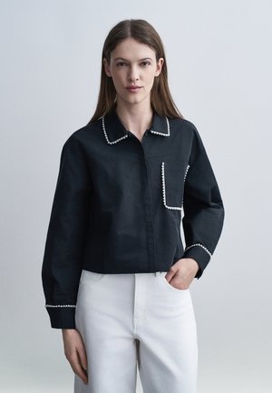 Γυναίκα με μακριά καστανά μαλλιά, ντυμένη με μαύρο πουκάμισο με κουμπιά και λευκή διακόσμηση από χάντρες, και λευκό παντελόνι, σε απλό ανοιχτόχρωμο φόντο.