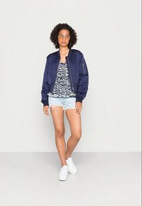 Casaco bomber azul marinho sobre uma blusa branca e azul com padrão, combinado com calções de ganga desgastados e ténis brancos. Conjunto casual moderno.