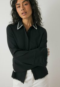 Black long-sleeve shirt with a collared neckline, featuring white scalloped trim on cuffs and hem, made from a smooth fabric.