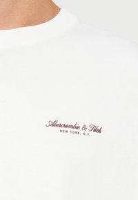 Bijela majica s okruglim izrezom, izrađena od pamuka, s malim maroon logotipom "Abercrombie & Fitch New York, N.Y." na lijevoj strani prsnog dijela.