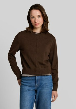 Junge Frau mit schulterlangem braunen Haar, die eine braune, geknöpfte Strickjacke und blaue Jeans vor einem schlichten grauen Hintergrund trägt.