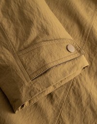 Beige stoff med krøllete tekstur har en trykknappdetalj på mansjetten og kontraststing langs kantene.