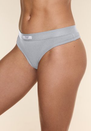 Ženski srednji del telesa, oblečen v svetlo sive spodnje perilo z etiketo Copenhagen Studios na enobarvni bež podlagi.