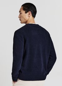 Ein Mann mit nach hinten frisiertem schwarzen Haar, der einen navyblauen Pullover und beige Hosen trägt, von hinten vor einem weißen Hintergrund betrachtet.