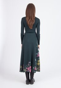 Donkergroen geribbeld sweater gecombineerd met een lange, gebreide rok met bloemenpatroon in roze en gele tinten, gedragen met kniehoge zwarte laarzen.