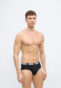 Καλά γυμνασμένος άνδρας με κοντά σγουρά μαλλιά, που φοράει μαύρα εσώρουχα Puma με λογότυπο στη μέση, στέκεται μπροστά σε απλό ανοιχτό γκρι φόντο.