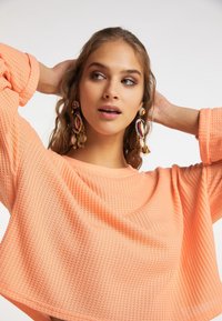 Pull court en tissu gaufré de couleur pêche vif, doté de manches larges. Le mannequin porte des boucles d'oreilles colorées avec des franges sur un fond neutre.