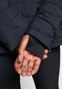 Giacca a vento blu navy con motivo trapuntato; polso di una camicia a maniche lunghe nera aderente visibile, che evidenzia il design a strati.