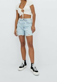 Bílý zkrácený top s uzlovým detailem, kombinovaný se světle modrými džínovými šortkami s třásněmi a černými vysokými teniskami s bílou podrážkou.