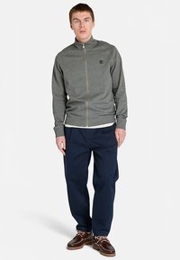 Grijze jas met ritssluiting van een gestructureerde stof, geribbelde manchetten en een kraag. Gecombineerd met marineblauwe broek en bruine schoenen. Eenvoudig, sportief ontwerp.